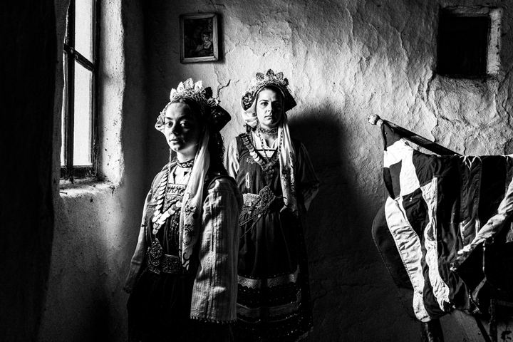 Two women standing by a window, wearing the local dresses of Paliouri in Evros, next to a greek flag