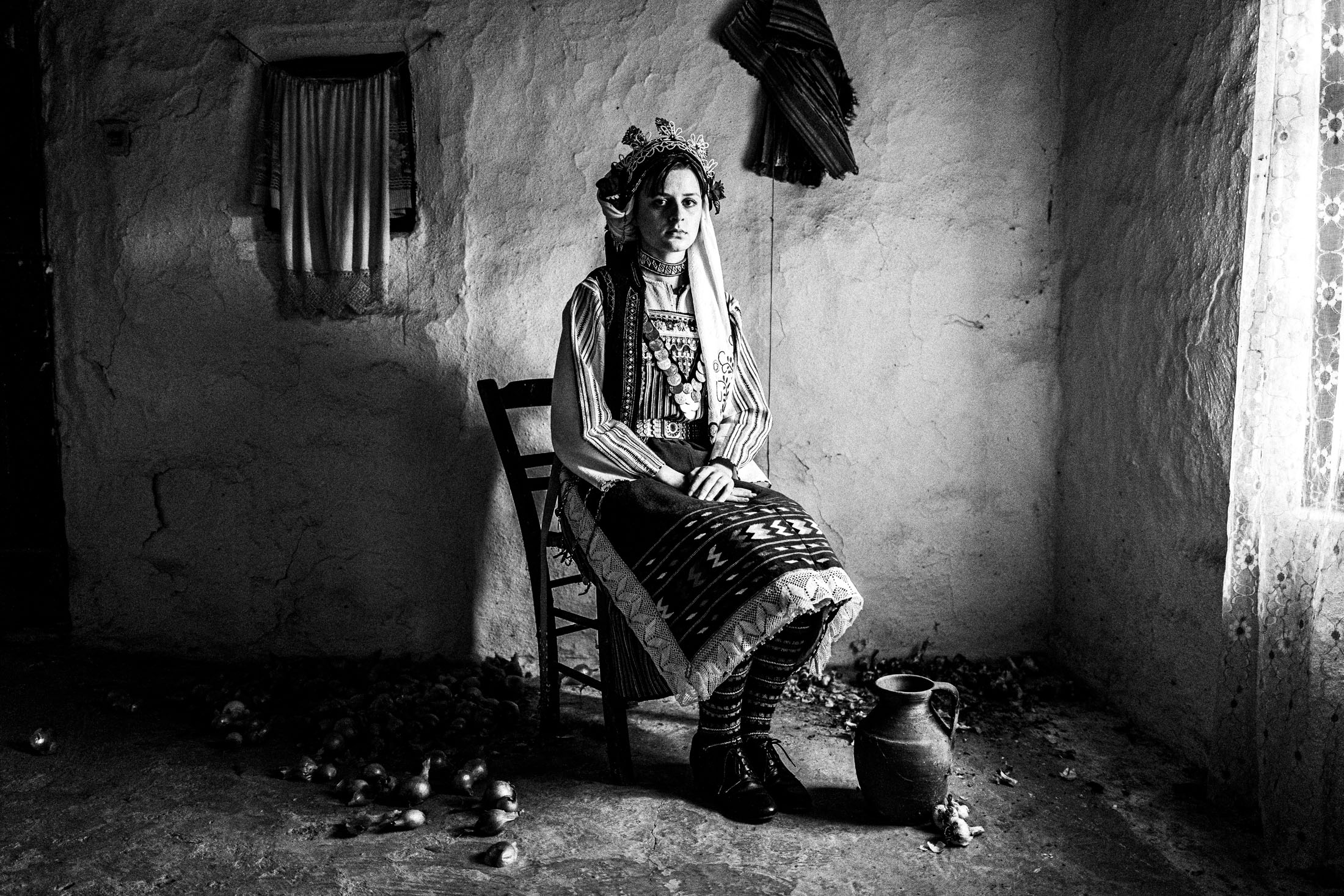 A woman wearing the local dress of Paliouri, sitting on a wooden chair by the window inside a local home.