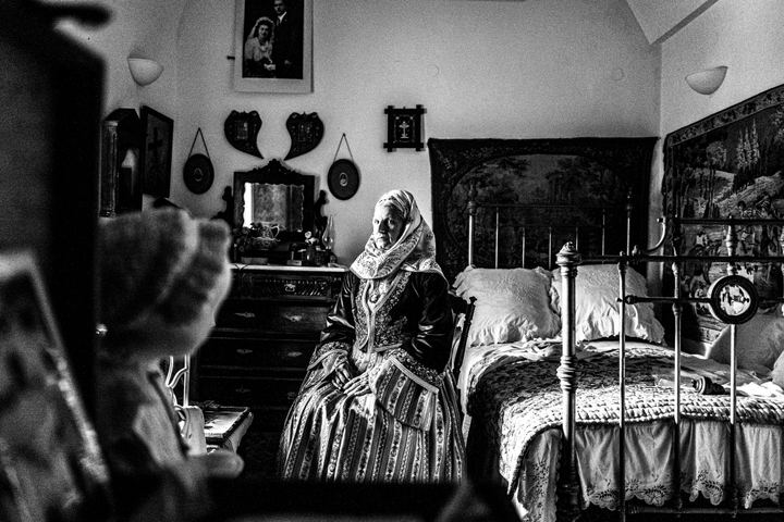 Woman wearing the local costume of Spetses, sitting by a bed inside a local traditional house