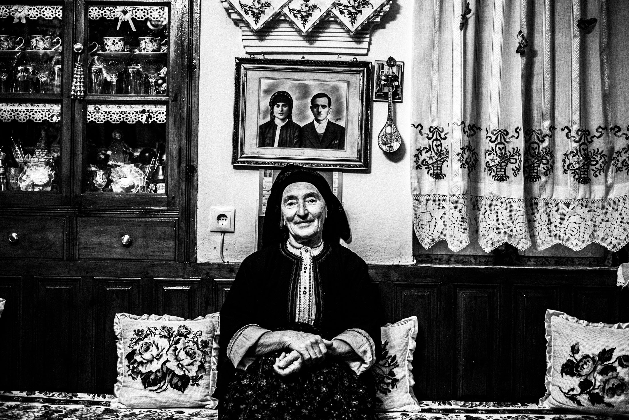 A woman in traditional clothes sits inside a local house with her hands crossed.