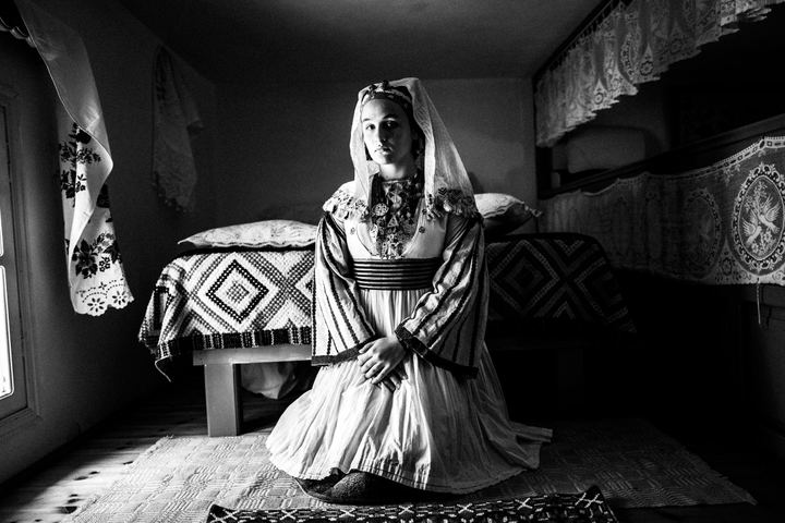 A young woman wearing the local costume of Tilos, sitting in front of a bed at a local house by a window