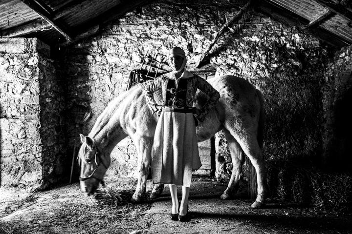 A teenage woman wearing the local costume of Pogoni inside a barn by a white horse in the village of Delvinaki, Epirus