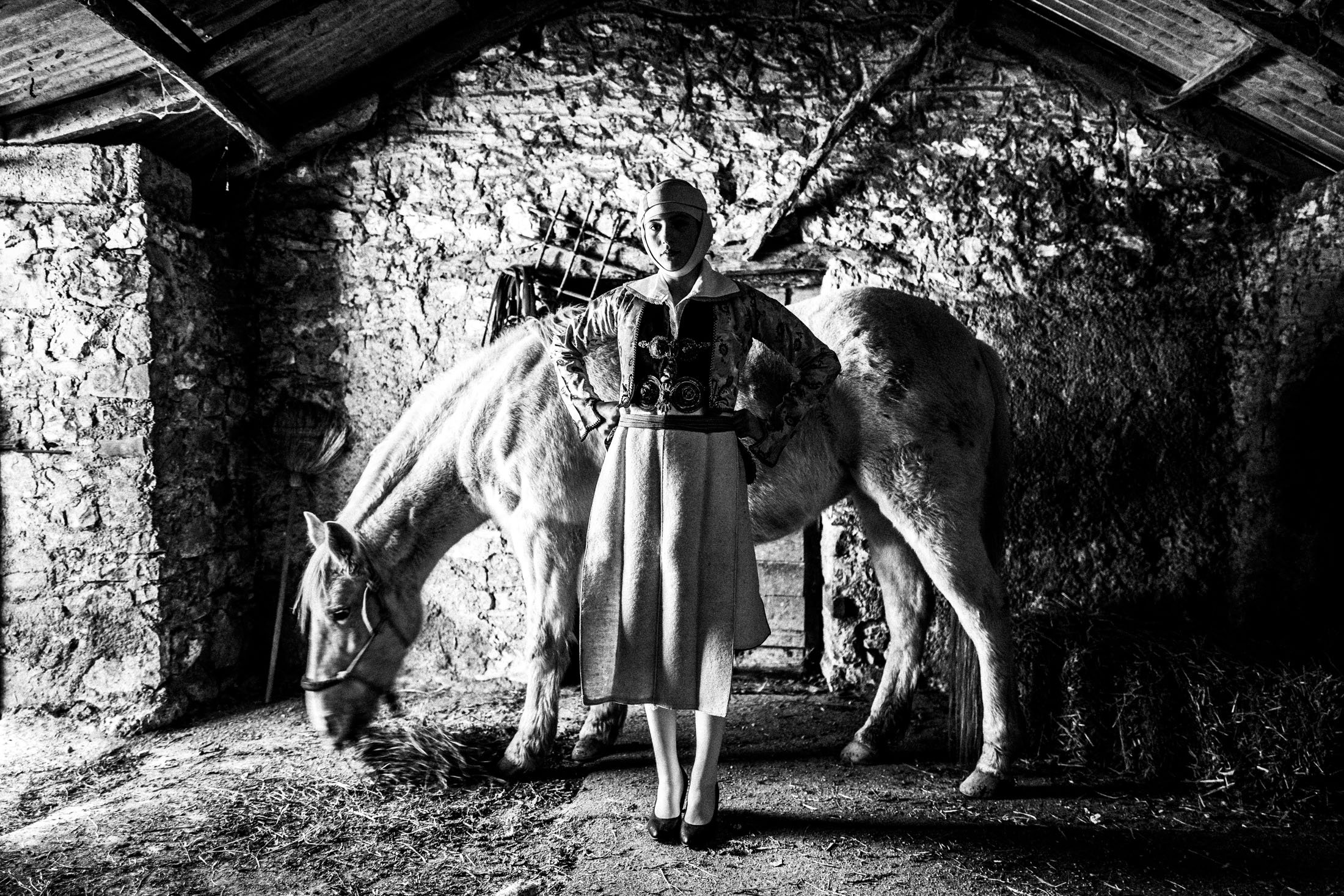 A teenage woman wearing the local costume of Pogoni inside a barn by a white horse in the village of Delvinaki, Epirus
