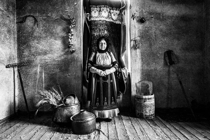 An elderly woman wearing the costume of Karagouna, standing in a room full of farmer's tools in Glinos, Trikala