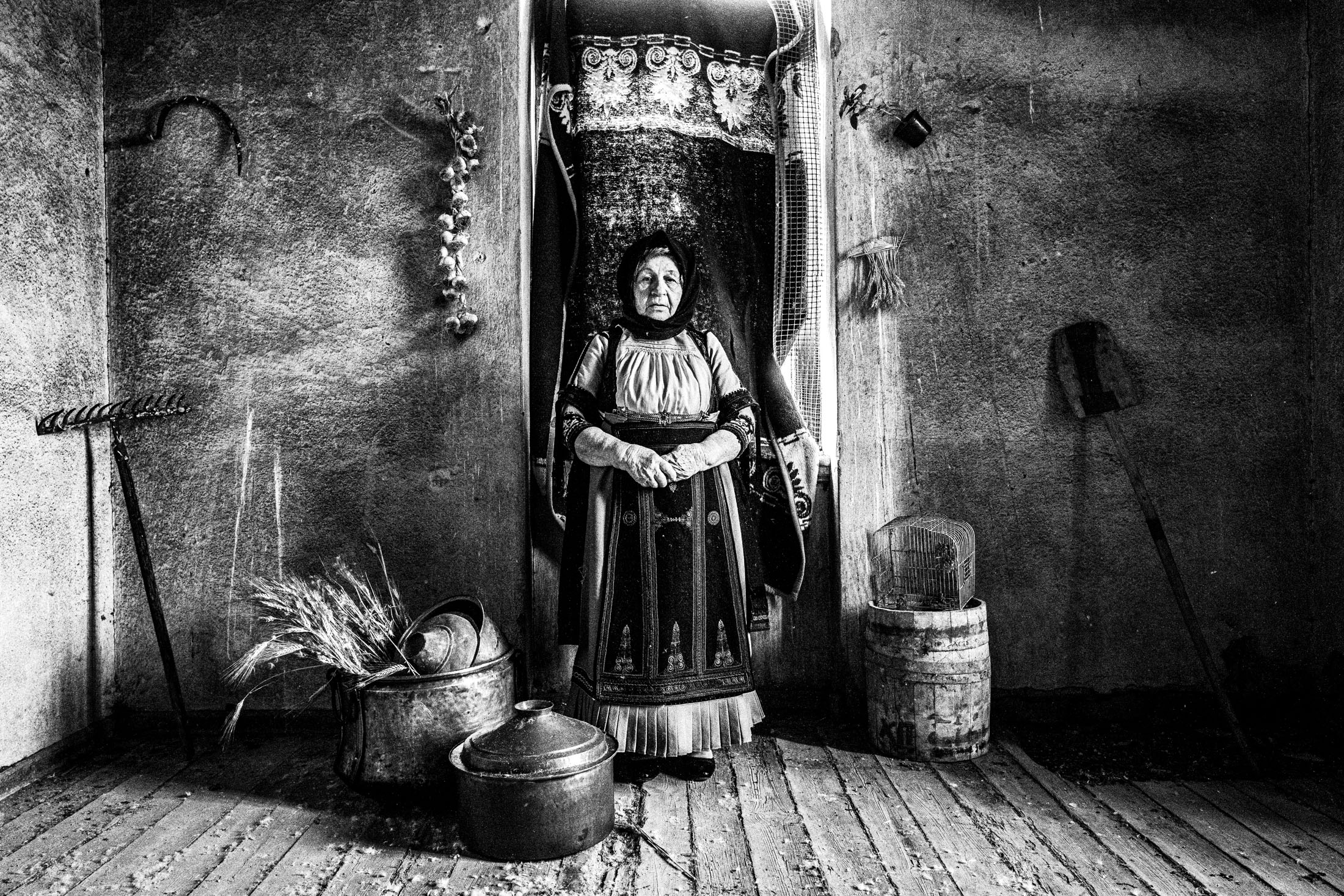 An elderly woman wearing the costume of Karagouna, standing in a room full of farmer's tools in Glinos, Trikala