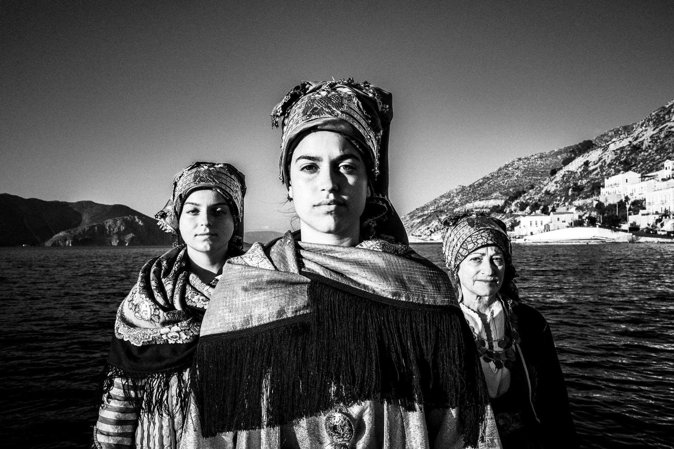 Three women wearing the local dresses of Symi, stand in formation on a boat before the sea