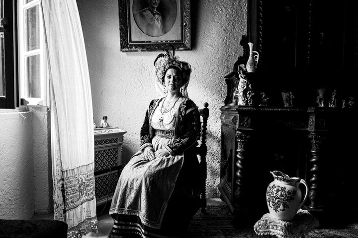 A woman wearing the costume of central Corfu, sitting on a wooden chair by a window inaide a local traditional house