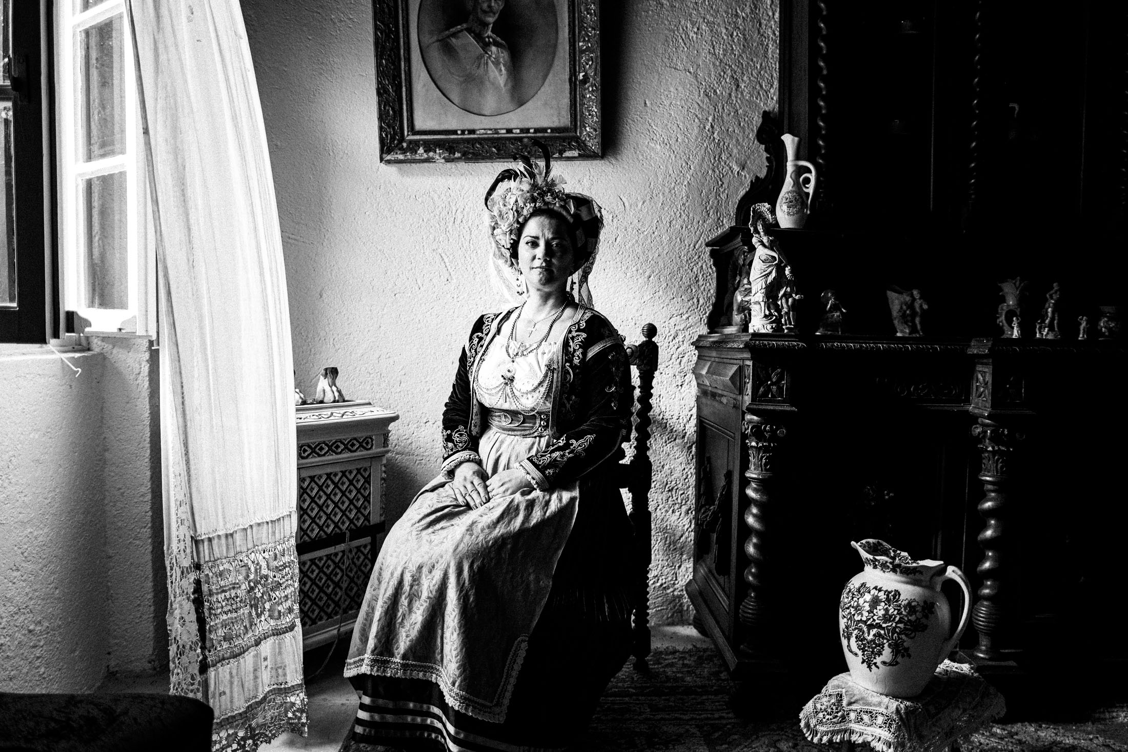 A woman wearing the costume of central Corfu, sitting on a wooden chair by a window inaide a local traditional house