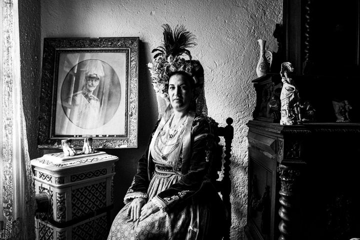 A woman wearing the costume of central Corfu, sitting on a wooden chair by a window inaide a local traditional house
