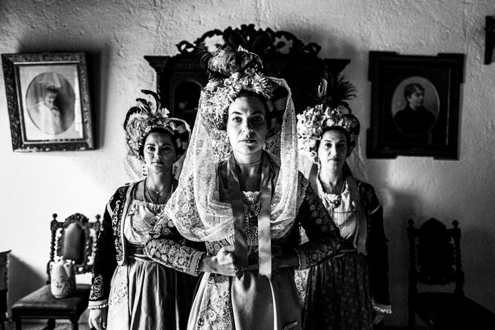 Three women wearing the costumes of central Corfu, standing in an arrow formation in front of a carved mirror inside a local traditional house