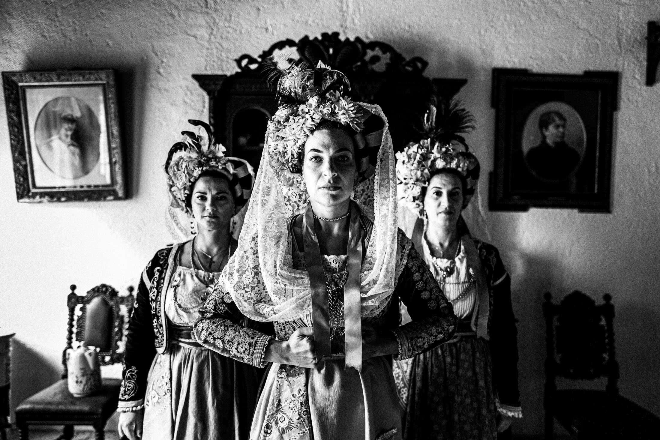 Three women wearing the costumes of central Corfu, standing in an arrow formation in front of a carved mirror inside a local traditional house