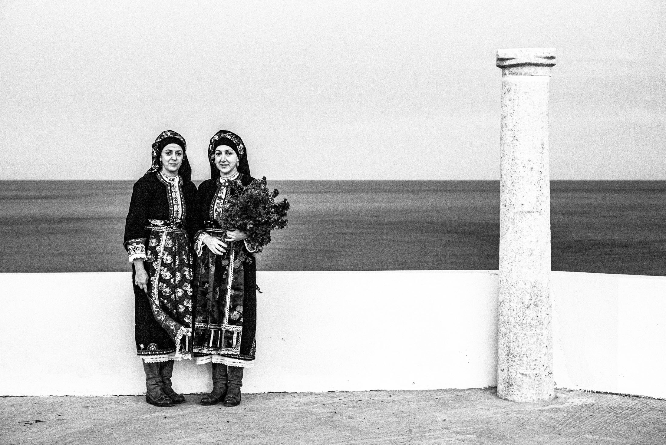 Two women wearing the local kavathe costumes of Olympos, by a column at Agios Minas, Karpathos.