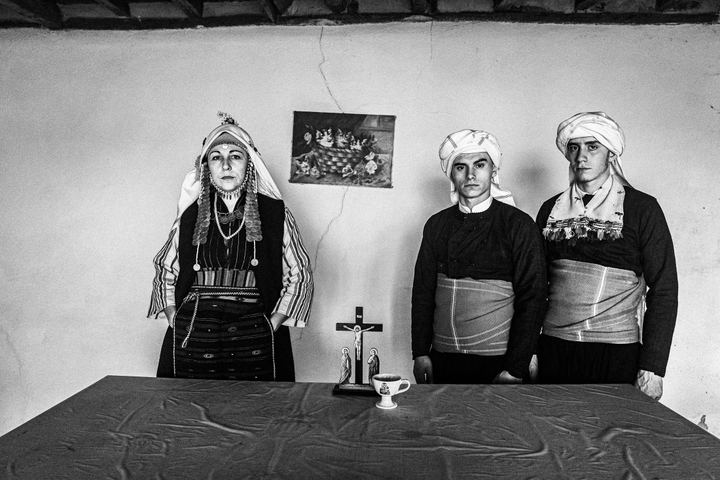 A woman and two men, dressed in the local costume of Ormenio, Evros, standing behind a table inside a local house in Mani, Thrace