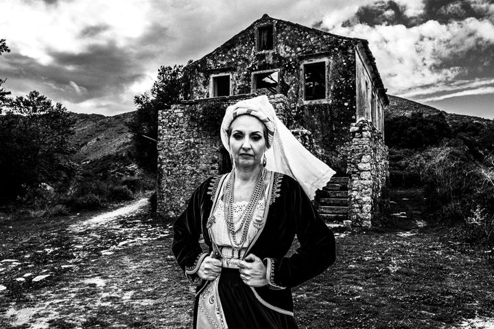 A woman wearing the local dress of Northern Corfu, standing in front of an old abandoned house in Palaea Perithea