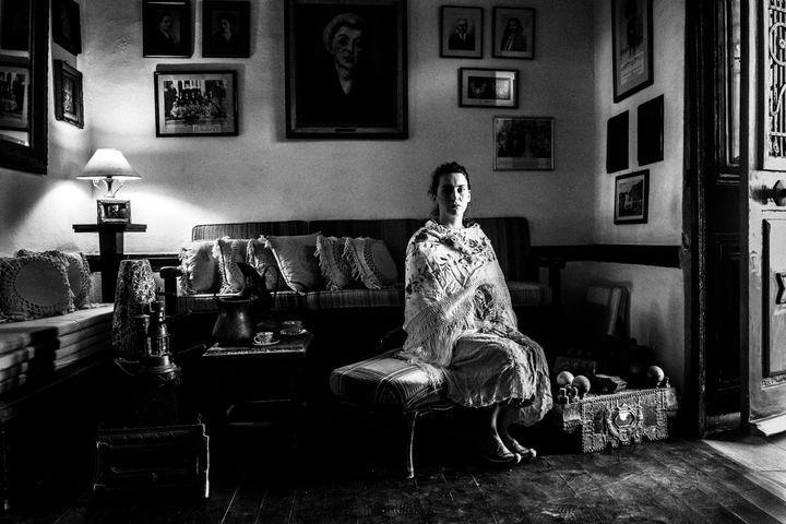 A woman wearing the local costume of Kastellorizo, sitting in a sofa, inside a local house by the window