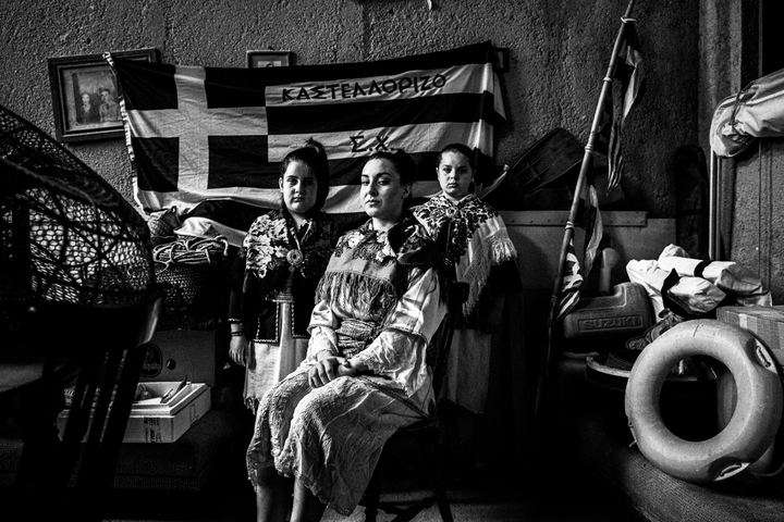 Three young women wearing the costumes of Kastellorizo, posing inside a local fisherman's house with a greek flag behind them