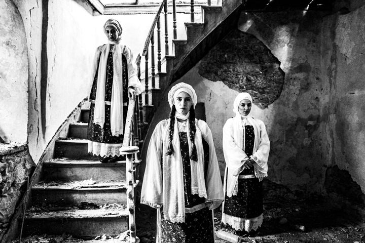Three women in the dresses of Vessa standing inside an old abandoned house by a staircase