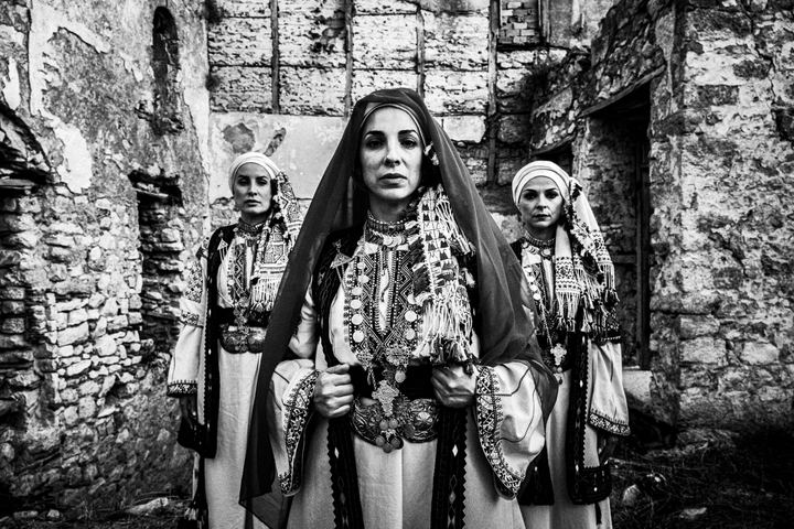 Three women wearing the local costumes of Kyparissia, Peloponnese, standing outside ruined local houses