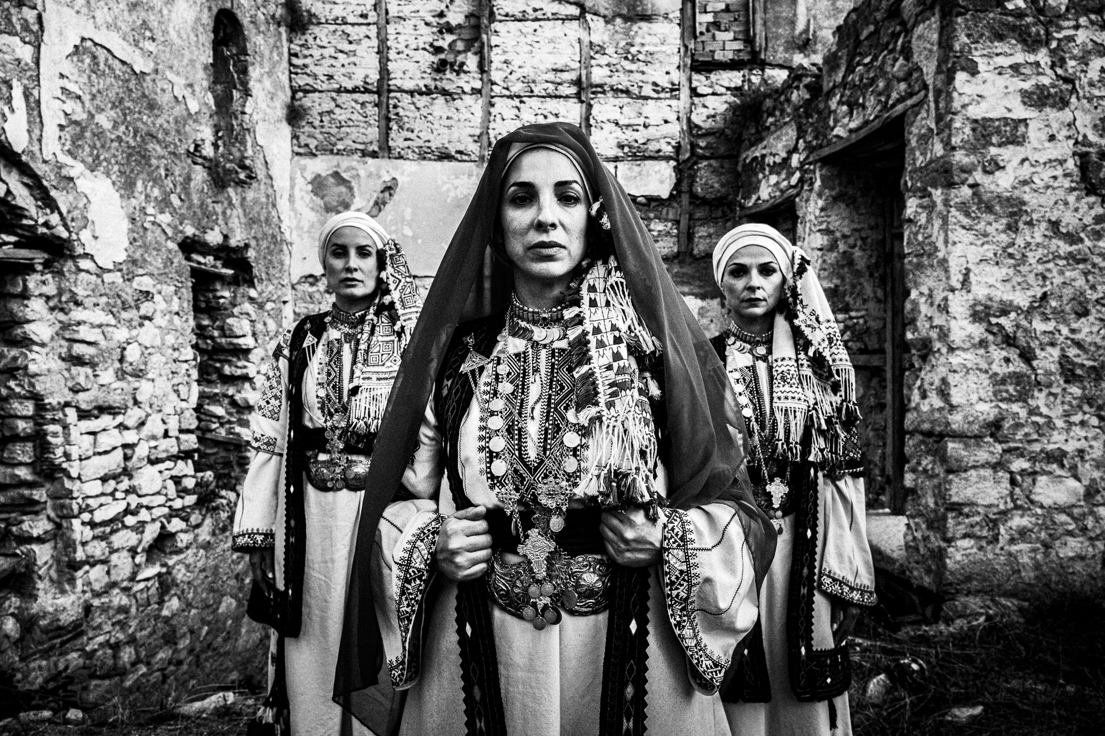 Three women wearing the local costumes of Kyparissia, Peloponnese, standing outside ruined local houses