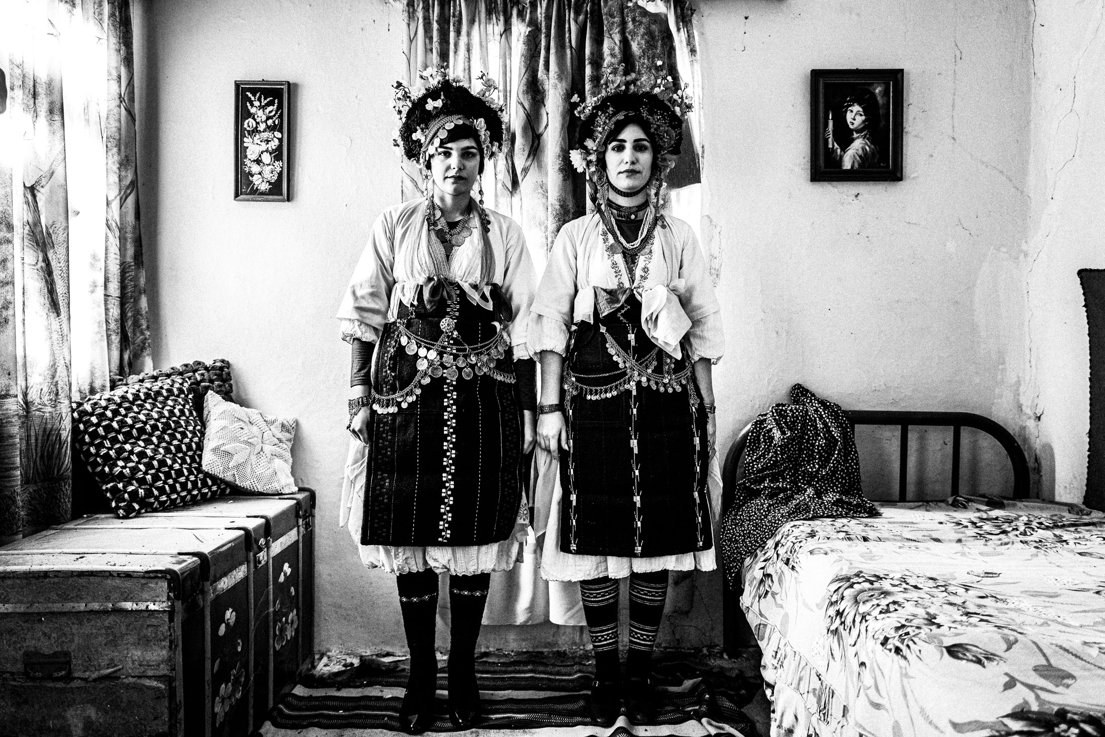 Two young women wearing the local Roumlouki Lazarines costumes, traditionally worn during Orthodox Easter, standing side-by-side inside a local house.