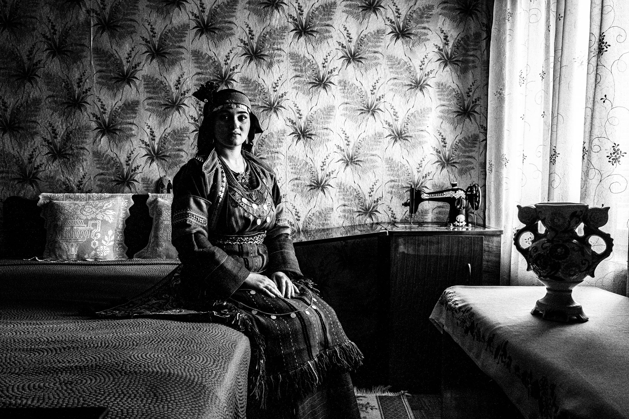 A woman wearing the local costume of Drymos in Thessaloniki, sitting on a bed by the window inside a local house