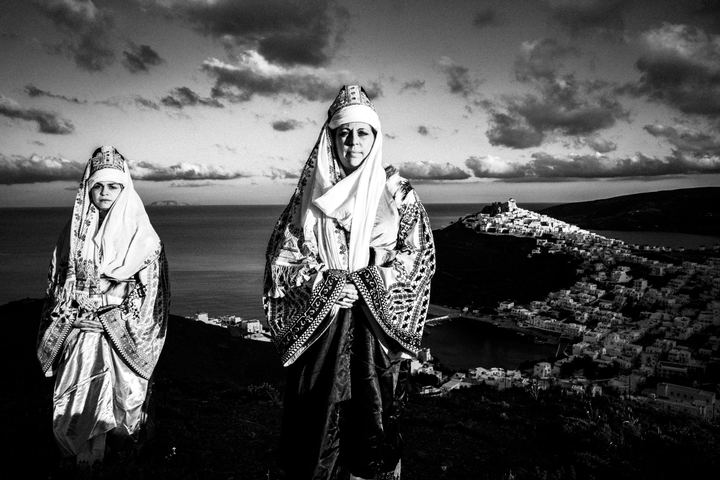 Two women wearing the local costume of Astypalaea, standing on a cliff overlooking the castle of Astypalaea