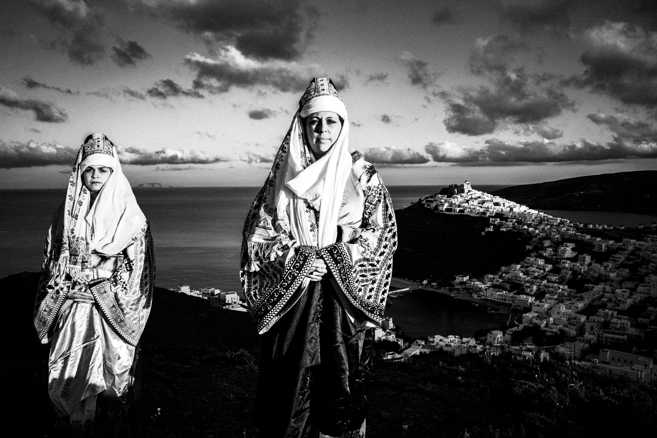 Two women wearing the local costume of Astypalaea, standing on a cliff overlooking the castle of Astypalaea