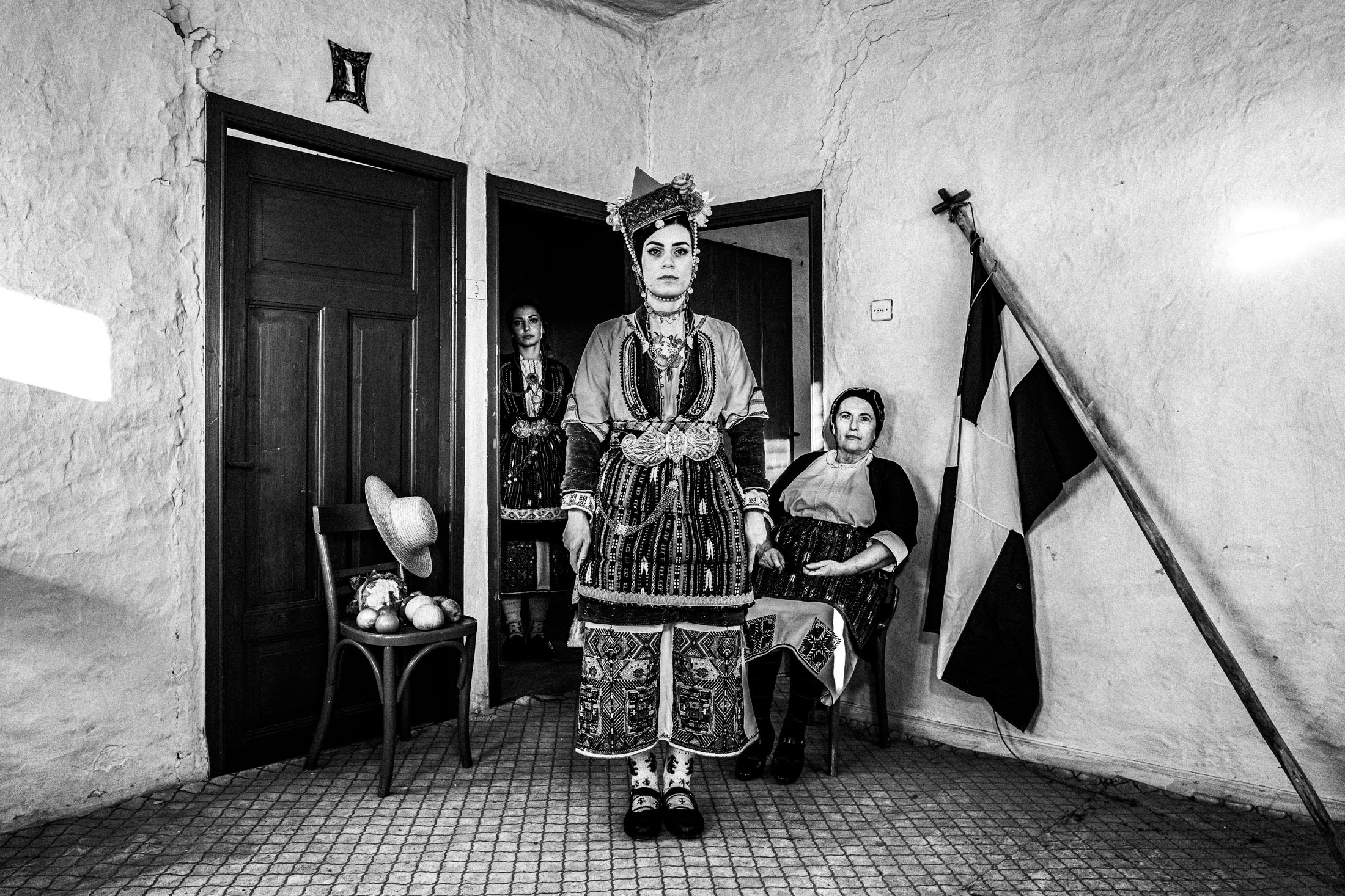 Three women, wearing the local costumes of the villages of the valley in Naoussa, Macedonia, posing in formation inside a local house, by a Greek flag