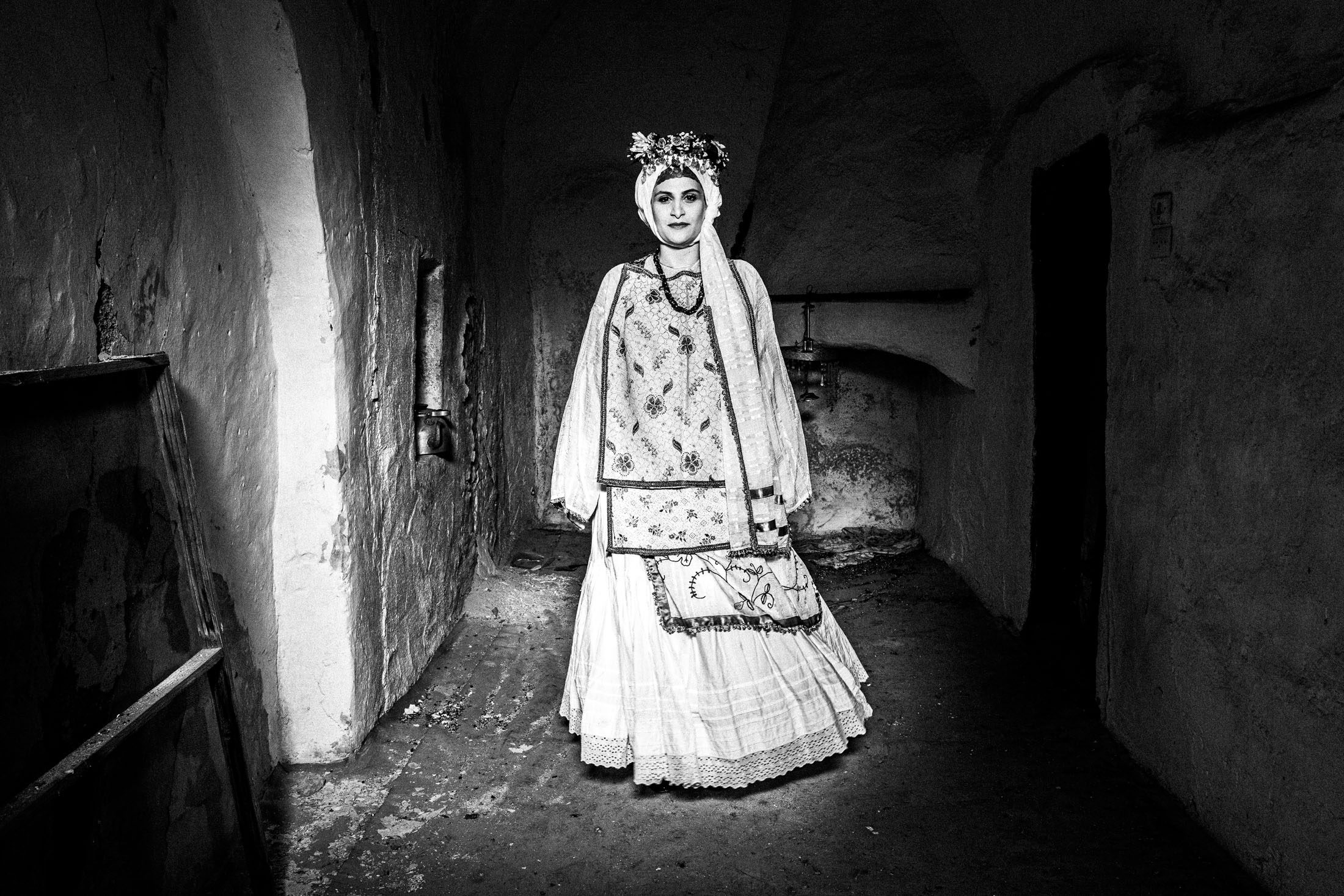 A woman wearing the local bridal dress of Agios Georgios Sikoussis, standing inside the ruins of a local house