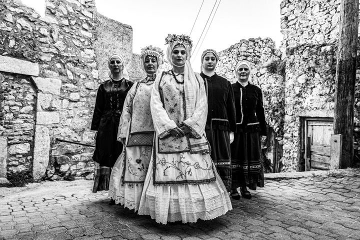 Five women wearing the local costumes of Agios Georgios Sykoussis, standing in formation inside the village