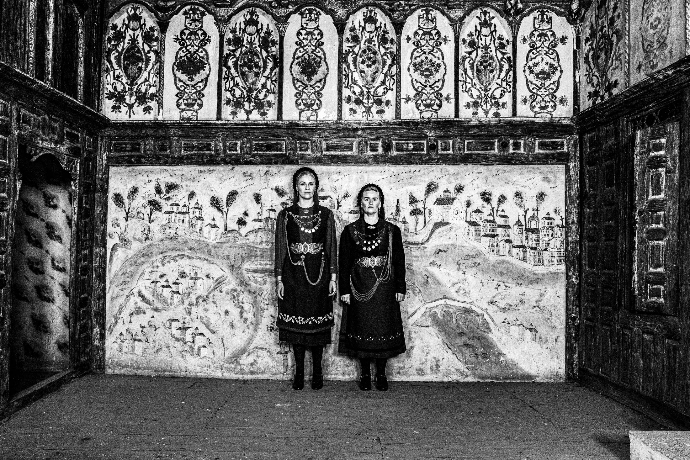 Two women wearing the kamvounia costumes, standing inside a traditional mansion in Siatista, before a painted wall