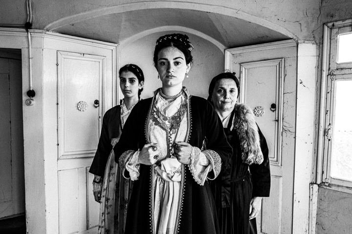 Three women wearing the urband costumes of Veria, standing in formation inside a local house.
