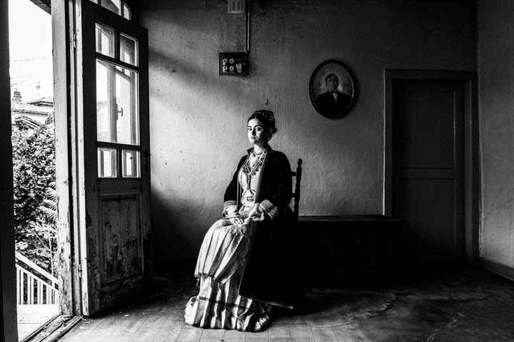 A woman wearing the urban costume of Veria, sitting inside a local house, by the door.
