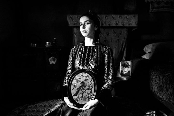 A young woman wearing the local costume of Agia Paraskevi (Kerasovo) in Epirus, sitting inside a local house, holding a framed embroidery of a bird
