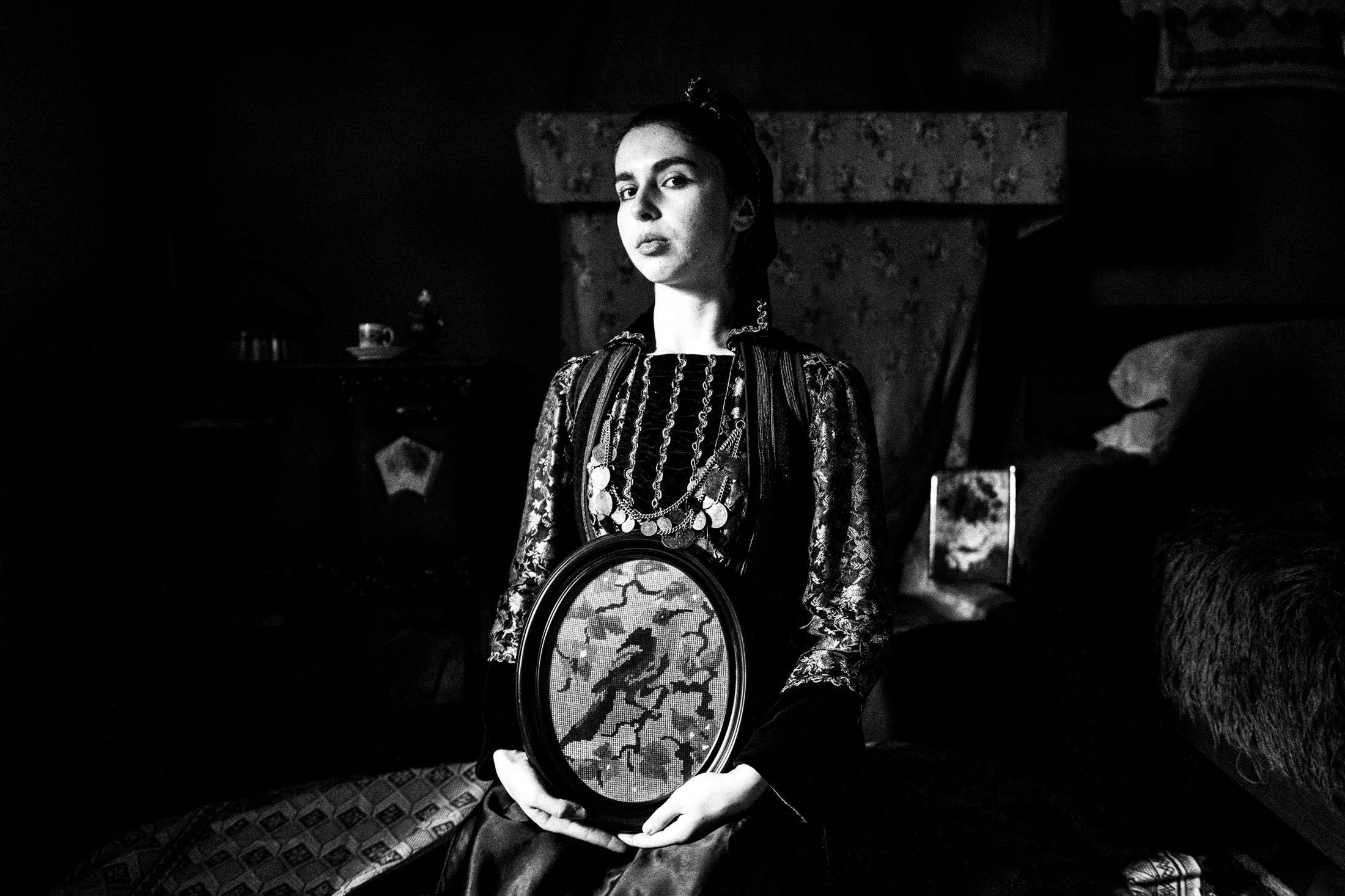 A young woman wearing the local costume of Agia Paraskevi (Kerasovo) in Epirus, sitting inside a local house, holding a framed embroidery of a bird