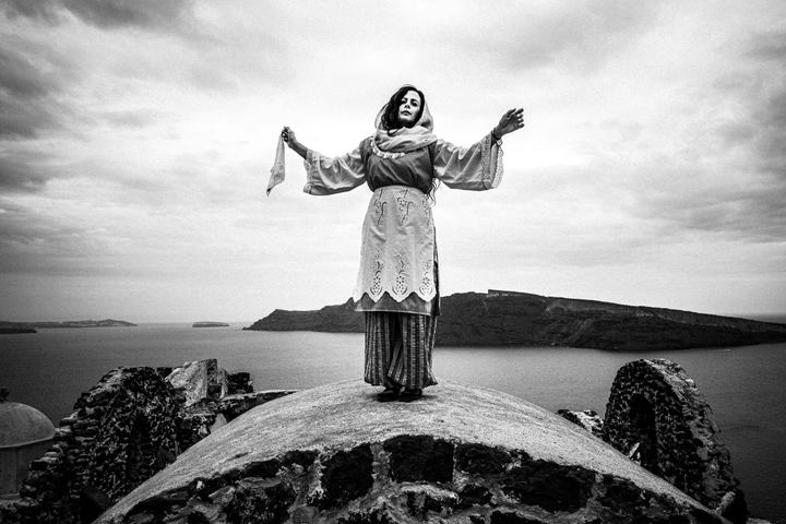 A woman wearing the local dress of Santorini, standing on a church dome in Oia, overlooking the Caldera.