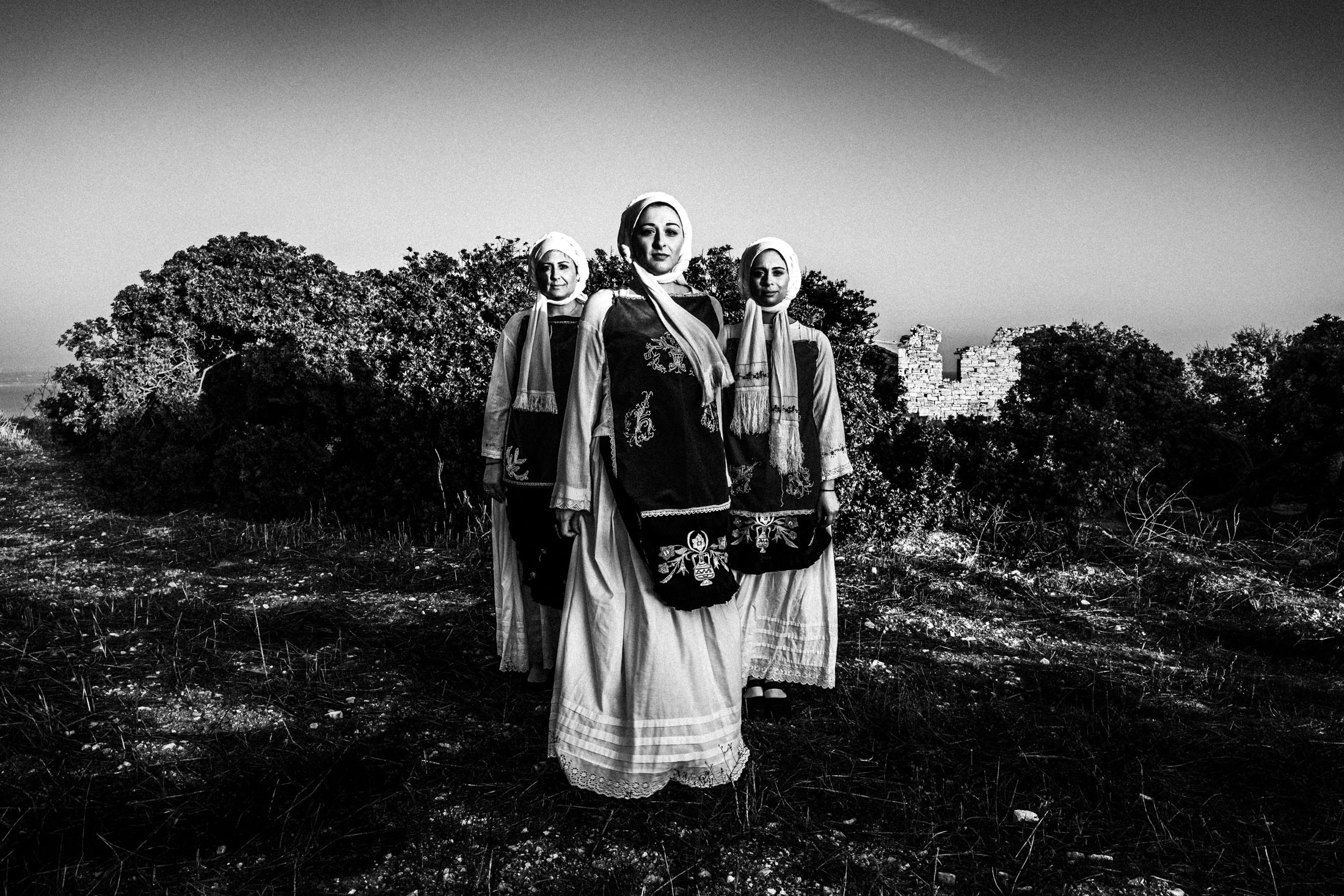 Three women standing in formation, wearing the local costumes of Nenita, Chios, in front of skinos trees and ancient ruins