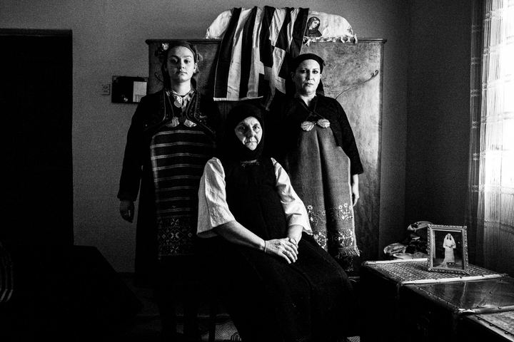 Three women wearing the costumes of Kostellides inside a house in Palatitsia, Imathia by a window, with a greek flag behind them.