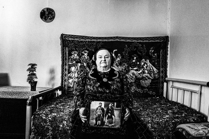 Woman wearing the local costume of Nea Vyssa in Evros, sitting on a bed, holding an old family photo.