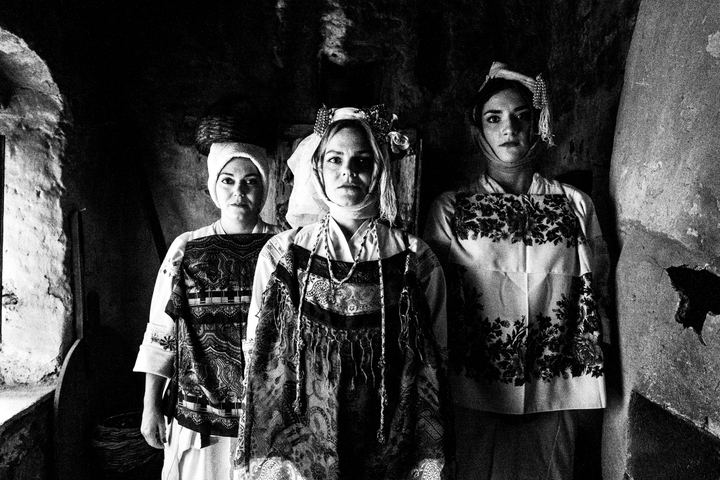 Three women sitting on a bed in an old house, wearing the local dresses of Pyrgi, Chios