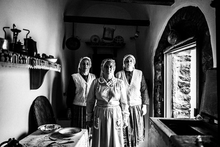 Three women wearing the local costumes of Mesta, standing in formation inside a local house