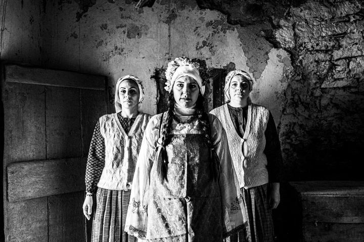 Three women wearing the local costumes of Mesta, standing in formation inside a local house