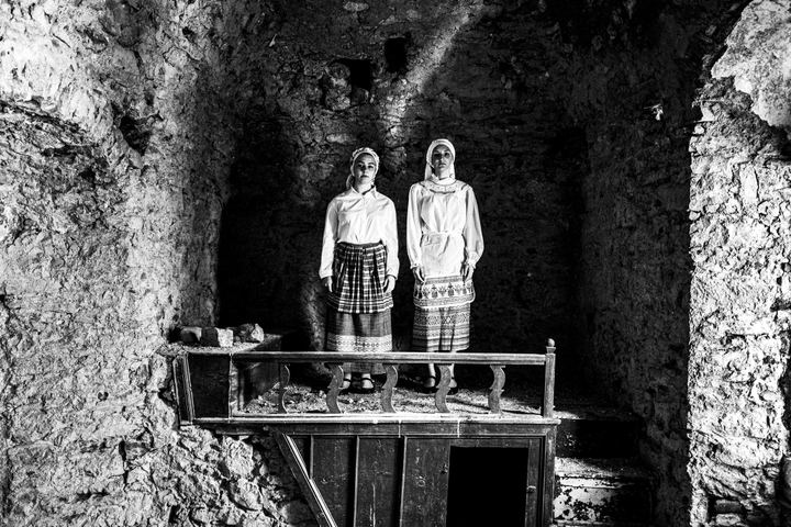 Two young women wearing the local costumes of Olympoi, standing on a wooden structure inside a local house