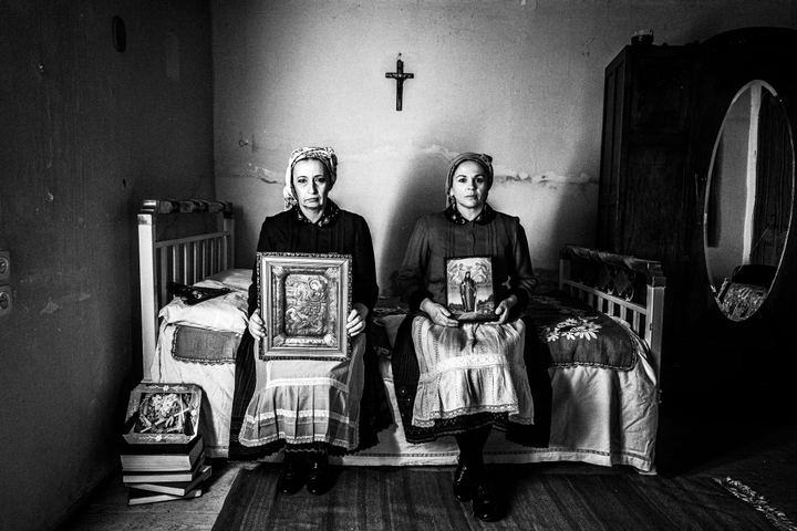 Two women wearing the local costumes of Divri, sitting on a bed holding orthodox icons inside a local house