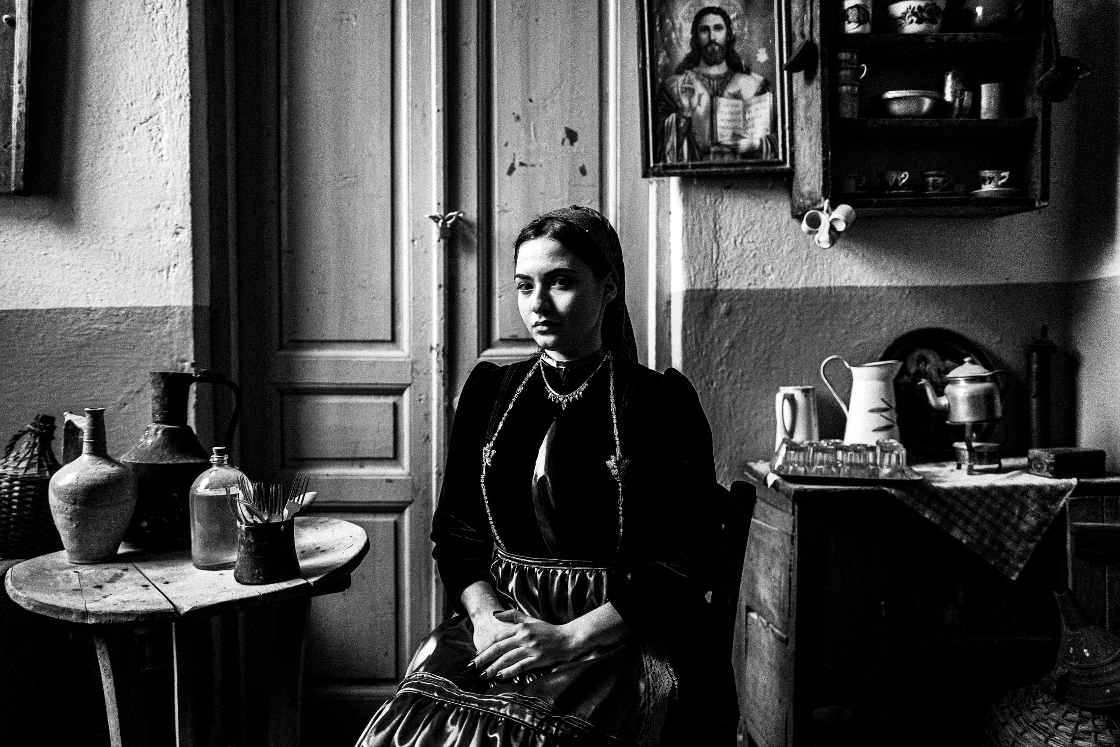 A woman in the dress of Polypotamos is sitting inside a local house with many props around her