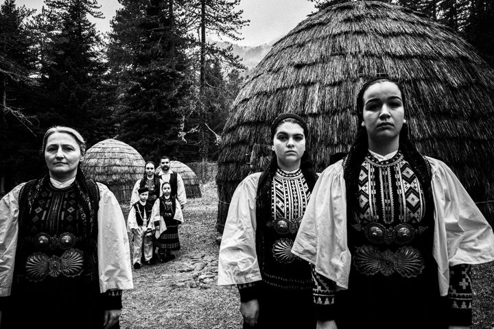 A group of people wearing the Sarakatsani nomadic group costumes, posing at Gyftokampos, Epiurs, by Sarakatsani traditional straw huts