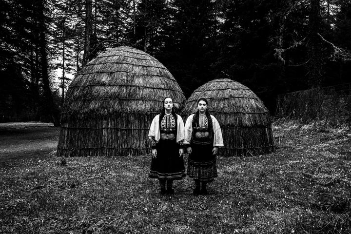 Two young women wearing the Sarakatsani nomadic group costumes, posing at Gyftokampos, Epiurs, by Sarakatsani traditional straw huts