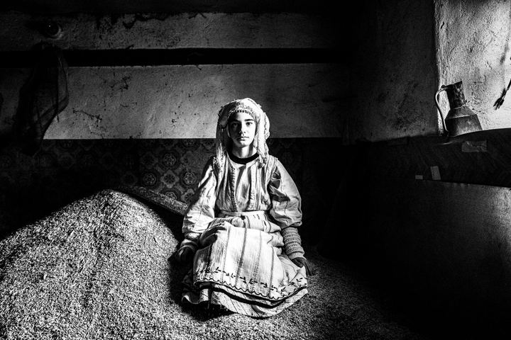 A young girl, wearing the local costume of Ermakia in Ptolemaida, sitting on a pile of grains by a window, inside a local abandoned house