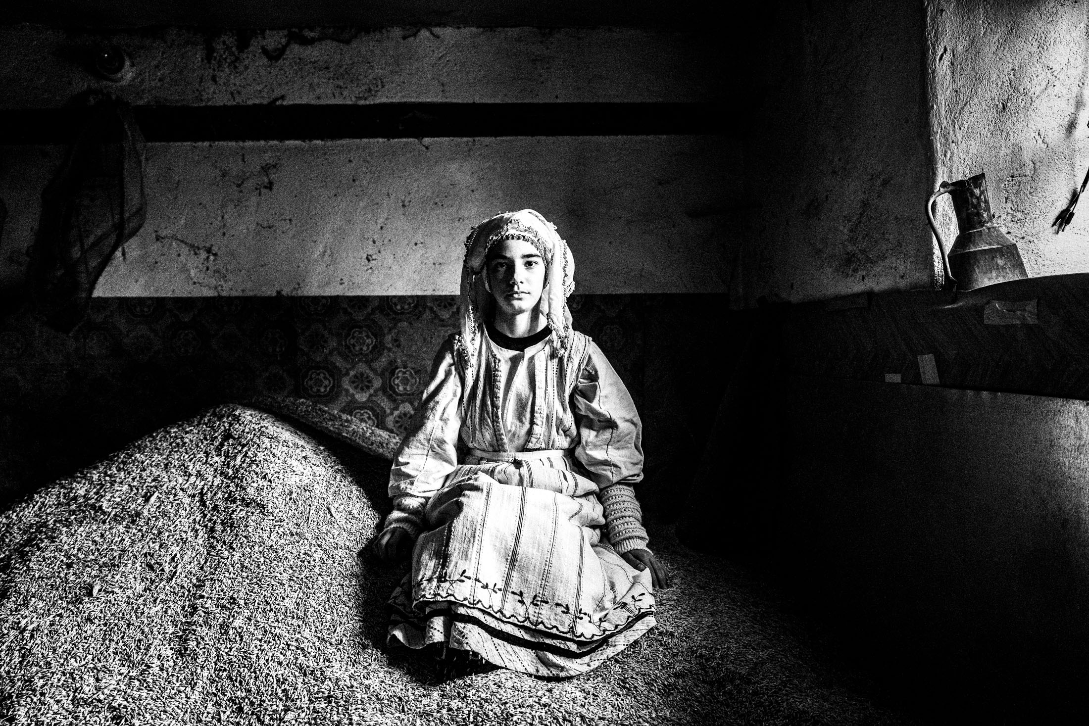 A young girl, wearing the local costume of Ermakia in Ptolemaida, sitting on a pile of grains by a window, inside a local abandoned house