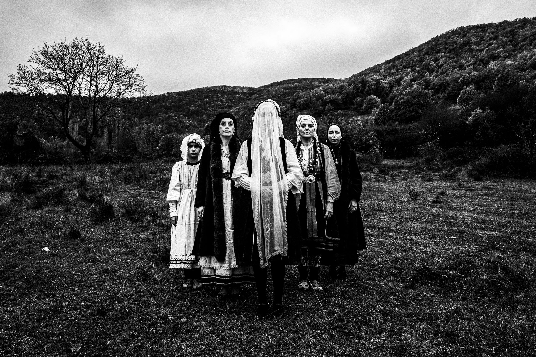 A group of women including a veiled bride, wearing the costumes of Ermakia, Eordaea, stand in formation on a mountain valley.